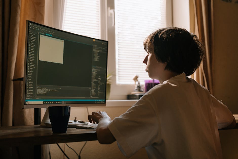 A woman with short dark hair sits at a wooden desk in a room illuminated by soft natural light from a window with blinds. She is focused on a large computer monitor displaying a coding interface with